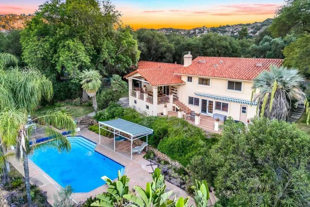 Aerial view of two-story Mediterranean-style home with terracotta tile roof and cream stucco exterior, featuring large rectangular swimming pool, covered patio, lush tropical landscaping with palm trees, and hillside canyon views at sunset