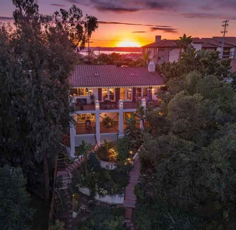 Twilight aerial view of multi-level Mediterranean-style home with terracotta tile roof and string lights illuminating wraparound porches, surrounded by mature trees and tropical landscaping, with sunset over water visible in background and neighboring home